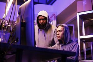Hackers identifying vulnerabilities in system to break into database and steal data. Asian and african american criminals in hoods hacking online server and cracking password on computer photo