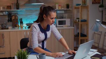 Concentrated freelance woman working overtime to finish a project from home kitchen. Busy focused employee using modern technology network wireless doing overtime for job reading typing, searching photo
