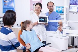 Dentist in dentistiry office showing tablet pc with jaw radiography to parent and little girl explaining treatment. Stomatologist explaining teeth diagnosis to mother of child in health clinic holding x-ray. photo
