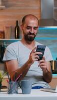 Freelancer taking break and searching on phone sitting on kitchen table working from home. Busy focused employee using modern technology network wireless doing overtime for job reading writing photo