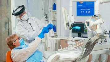 Nurse in coverall helping doctor with patient dental x-ray during examination in coronavirus pandemic. Medical team wearing face shield mask, gloves, explaining radiography using monitor display photo