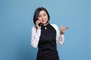 Cafe waitress smiling while managing conversation with client on mobile phone. Joyful carefree young woman receptionist wearing professional uniform chatting on smartphone photo