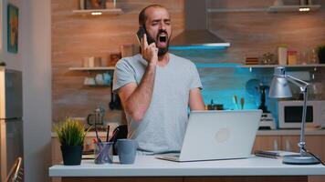 Tired freelancer talking on phone yawning sitting at kitchen desk in on laptop late at night. Busy employee using modern technology network wireless doing overtime for job reading writing, searching photo