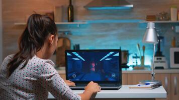 Woman gamer sitting at home in kitchen testing new game on professional laptop. Tired player gaming online video games on her personal computer with modern technology network wireless photo