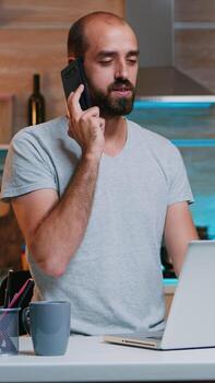 Bored man in t-shirt yawning while talking on smartphone working at home with laptop. Busy focused employee using modern technology network wireless doing overtime for job reading writing, searching photo
