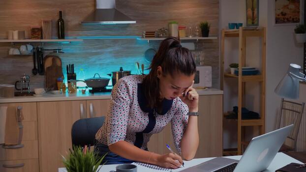 Businesswoman writing notes while working late on computer in home kitchen. Busy focused employee using modern technology network wireless doing overtime for job reading typing, searching photo