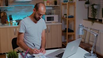 Businessman writing notes while working late on computer in home kitchen. Busy focused employee using modern technology network wireless doing overtime for job reading typing, searching photo