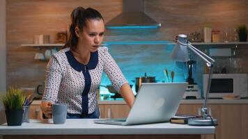 Remote employee working late night at deadline sitting on desk in home kitchen. Busy focused businesswoman using modern technology network wireless doing overtime for job reading writing, searching photo