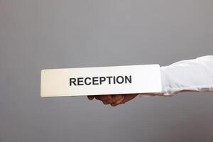 Hotel administrator hand holding reception stainless steel plate closeup. Hospitality service receptionist arm showing tabletop metal name sign close view in studio on gray background photo