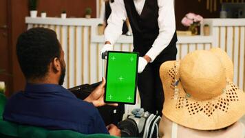 Guests holding tablet with greenscreen in lounge area lobby, using isolated display together in reception. Tourists couple dressed in summer clothes looking at copyspace template. Tripod shot. photo