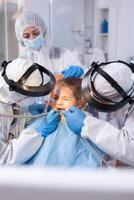 Mother comforting ittle girl with painfull face expression in the course of dental treatment. Stomatology team wearing ppe suit during covid19 doing procedure on child teeth. photo