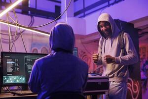 Cheerful internet scammers discussing phishing scam and planning online fraud. Smiling african american hacker explaining data breach idea to criminal in abandoned warehouse photo