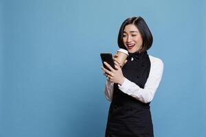 Cheerful asian waitress laughing while drinking takeaway coffee and scrolling social media on smartphone. Smiling woman receptionist enjoying beverage to go and using mobile phone photo