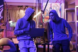 Twp hackers breaching cybersecurity system to gain illegal access. Man in hood showing computer virus code on laptop to malicious software developer and asking for advice photo