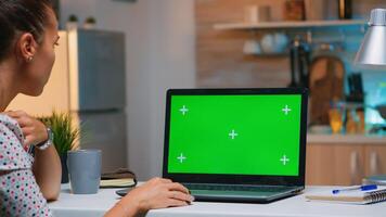 Freelancer looking at green screen monitor sitting at home in kitchen. Business woman watching desktop laptop display with green mockup, chroma key, during night time working overtime. photo