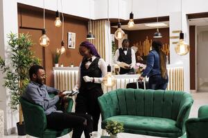 Smiling friendly hotel waitress accepting contactless NFC mobile payment from guest resting in lobby. Happy african american tourist using smartphone to pay for room during check out process photo