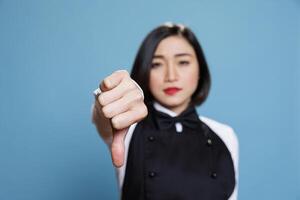 Young asian woman wearing waitress uniform showcasing disapproval with thumb down gesture portrait. Restaurant receptionist showing service negative feedback concept and looking at camera photo