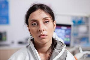 Exhausted dentist wearing protective gear agasint coronavirus looking at camera. Tired medical specialist in dentistry cabinet wearing safety equipment during global health crisis. photo