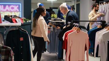 Diverse clients shopping in retail store, looking for black friday discounts. Customers browsing through clothing items on hangers and racks in fashion outlet, exploring seasonal sales. photo