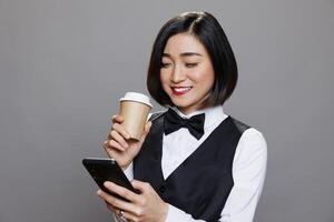 Smiling asian woman dressed in waitress uniform holding coffee paper mug and browsing internet on smartphone. Receptionist enjoying tea and engaging with social media networks on mobiel phone photo