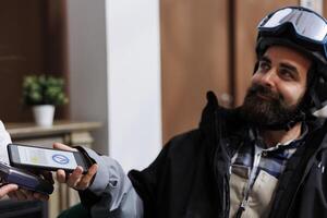 In ski resort lounge area, caucasian man pays with cell phone while staff assists with pos machine. Close-up view of male traveler wearing wintersport outfit using smart device for nfc payment. photo