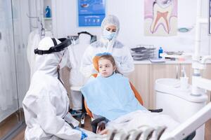 Dentist in coverall talking with kid before dentistiry examination with face mask. Stomatologist during covid19 wearing ppe suit doing teeth procedure of child sitting on chair. photo