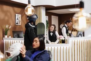 Young man wearing helmet and holding snowboard arrives at hotel reception greeted by friendly staff. Guests in snow gear going for wintersport activity at luxury ski mountain resort. photo