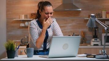 Woman yawning while working remotly from home late at night in the kitchen. Busy exhausted employee using modern technology network wireless doing overtime for job reading typing, searching photo