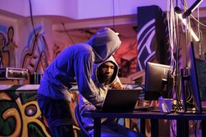 Criminals hacking on laptop together and planning ddos attack on website. Two hackers in hoods checking malcious software code on portable computer in abandoned warehouse photo