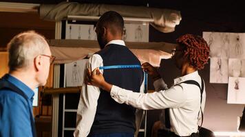 Tailors team taking measurements for client, working in atelier to create custom made tailored suit. Couturier and assistant measuring customer to design perfect fit clothes. Handheld shot. photo