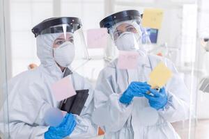 Dentists discussing about patient teeth dressed in ppe suit during covid-19. Medical team in stomatology office wearing coverall in dental office writing ideas on sticky notes. photo