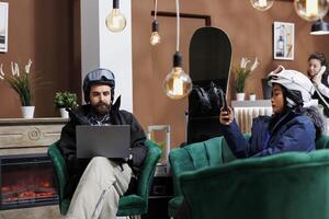 Two travelers in snow clothing using their technology devices while waiting for check-in process at luxury winter resort. Couple holding their laptop and phone while seated in ski hotel reception. photo