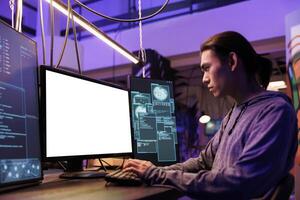 Hacker spreading malware and doing illegal activities on computer with white screen. Young asian criminal breaking law while programming malicious malware in abandoned warehouse photo