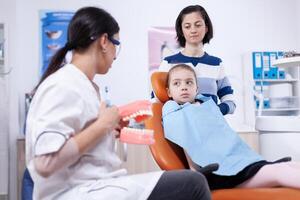 Stomatologist in the course of teeth brushing artificial jaw in the course of child check up. Little girl and mother listening stomatolog talking about tooth hygine in dentistiry clinic holding jaw model. photo