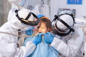 Woman dentist doctor treating little girl tooth with pain face expression. Stomatology team wearing ppe suit during covid19 doing procedure on child teeth. photo