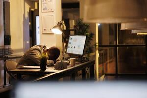 Tired drained entrepreneur sleeping on desk in startup office, workaholic employee falling asleep after finishing financial report. Exhausted african american worker with burnout syndrome resting at work. photo