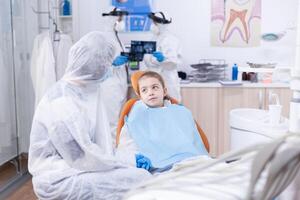 Stomatologist during covid19 wearing ppe suit doing teeth procedure of child sitting on chair. photo