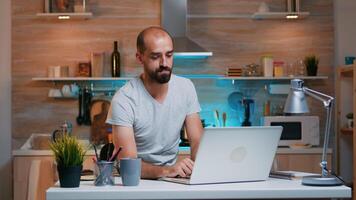 Remote man entrepreneur having video conference with colleagues using laptop sitting in the kitchen late at night. Busy employee using modern technology network wireless reading writing, searching photo