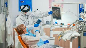 Stomatologist in protective suit reviewing x-ray of tooth with senior patient explaining treatment using tablet in covisd-19 pandemic. Medical team wearing face shield, coverall, mask and gloves. photo