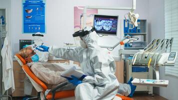 Dentist nurse in potective suit controlling patient temperature before dental examination during coronavirus pandemic. Concept of new normal dentist visit in coronavirus outbreak wearing protective suit and face shield photo