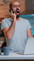 Tired freelancer talking on phone yawning sitting at kitchen desk in on laptop late at night. Busy employee using modern technology network wireless doing overtime for job reading writing, searching photo