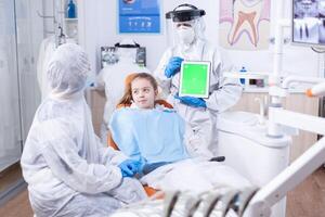 Tablet pc with green screen used by dentist in ppe suit during consultation of little girl. Medical specialist in oral hygine holding tablet pc with copy space available during global pandemic with covid19. photo