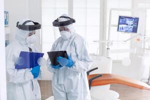 Professional dentists using tablet pc dressed in ppe suit as safety precaution for covid19. Stomatology team in dental office wearing protective suit agasint contagious coronavirus during global pandemic. photo