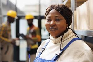 Smiling african american woman working in industrial warehouse portrait. Shipment manager with positive facial expression standing near parcels shelf and looking at camera close up photo