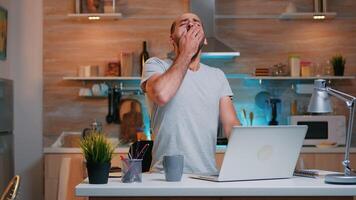 Tired sleepy man yawning, working at home desk in the kitchen. Busy overwork focused employee using modern technology network wireless doing overtime for job reading writing, searching photo
