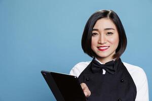 Smiling cafe waitress holding digital tablet at work and looking at camera with cheerful facial expression. Joyful cafe woman worker using portable gadget portrait on blue background photo