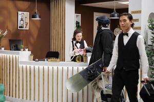 Female receptionist at front desk assists man with snowboarding equipment with accommodation reservation at winter resort. Professional bellboy taking luggage of male guest towards booked hotel room. photo