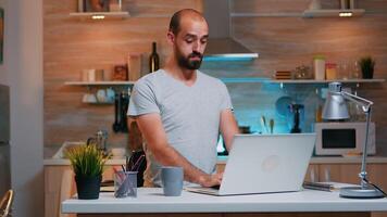 Exhausted freelancer yawning sitting at kitchen desk in front of laptop late at night. Busy focused employee using modern technology network wireless doing overtime for job reading writing, searching photo