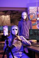 Scammers in anonymous masks streaming threat online and demand victim for payment. Hackers with hidden identity filming ransom message on mobile phone in abandoned warehouse photo