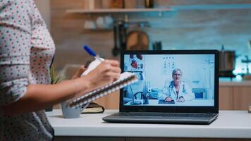 Lady writing notes during online medical consultation listening woman doctor sitting in front of laptop in the kitchen. Sick person discussing during video conference about symptoms and treatment. photo